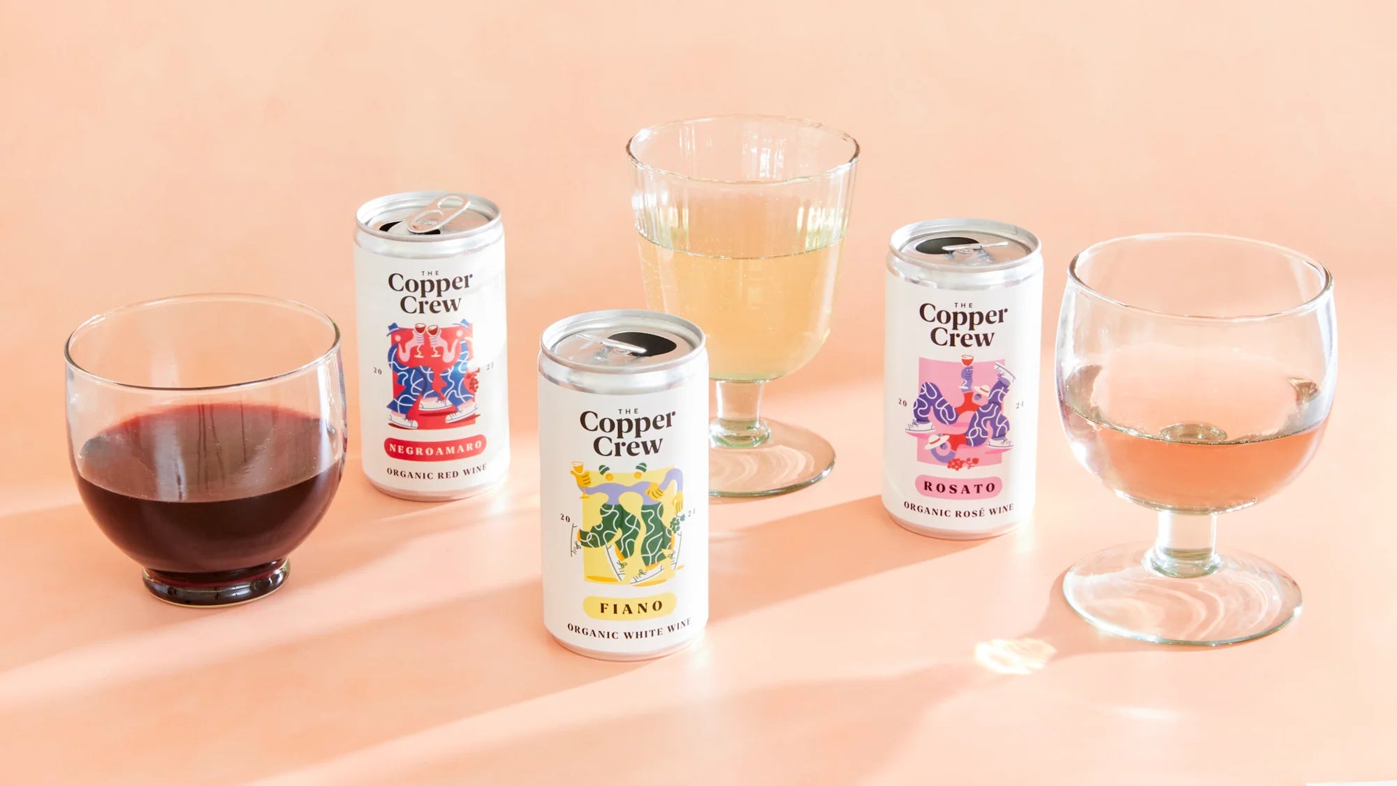 The Copper Crew canned wine collection with glasses of organic red, white, and rosé wine displayed on a peach background