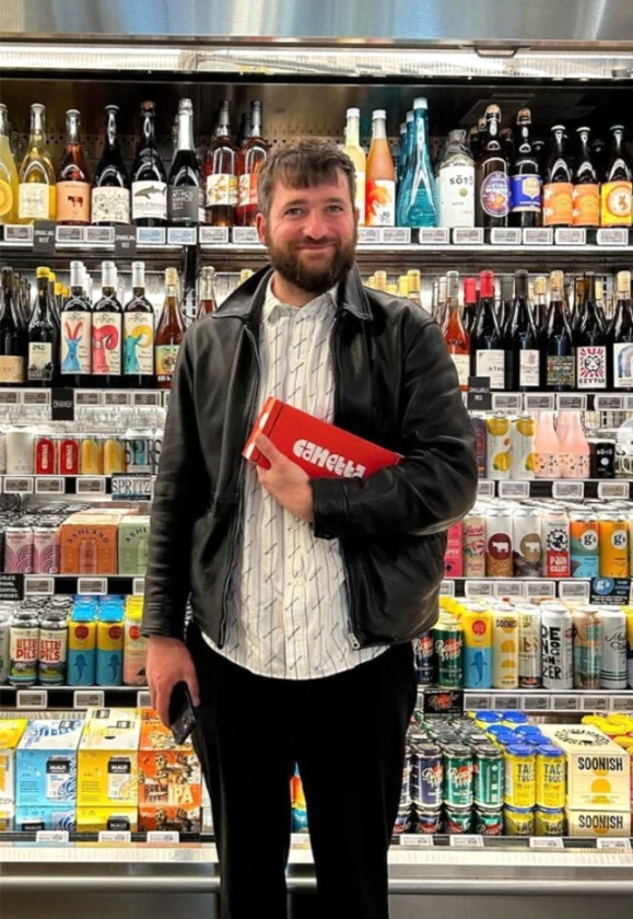 Luca Pronzato, founder of Canetta, holding a pack of canned wine in front of a wine fridge, highlighting the brand’s innovative natural wine approach.