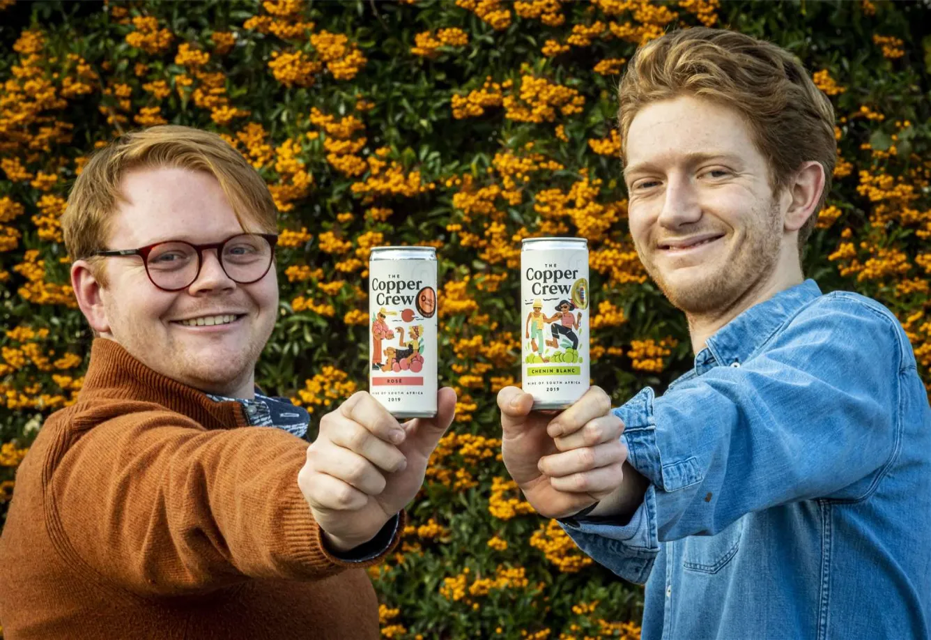 The Copper Crew founders Oli Purnell and Theo Gough holding their canned wines in front of a floral background